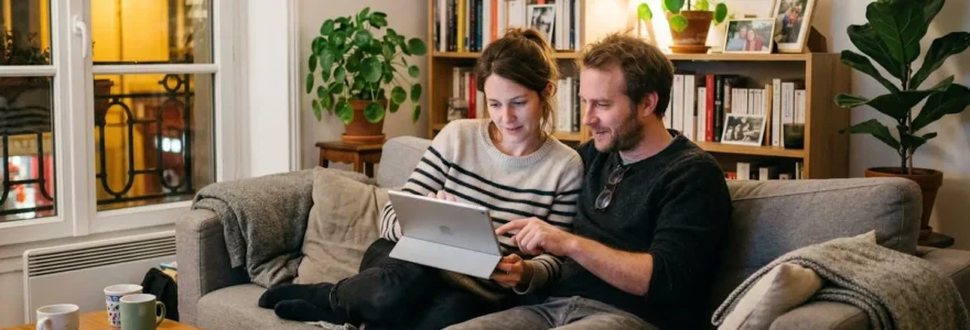 Un couple d'une trentaine d'années assis sur un canapé moderne consulte ensemble une tablette dans un salon lumineux